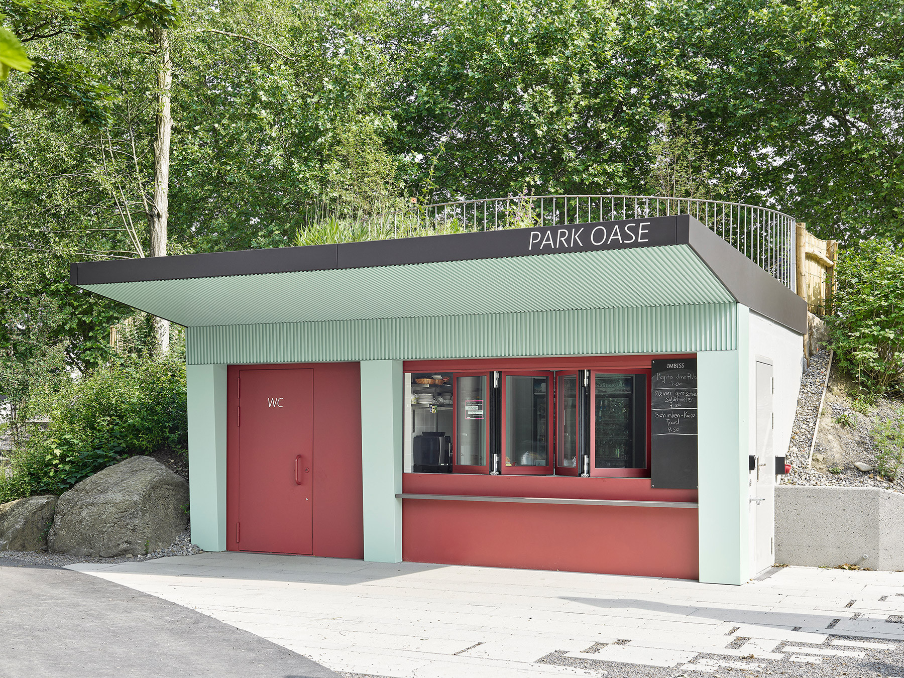 Buvette „Park Oase“ im Kyburgpark Sursee mit grün beschichteten Stahlblechen, roten Türen und Fenstern, grossem Stahlvordach und Dachterrasse