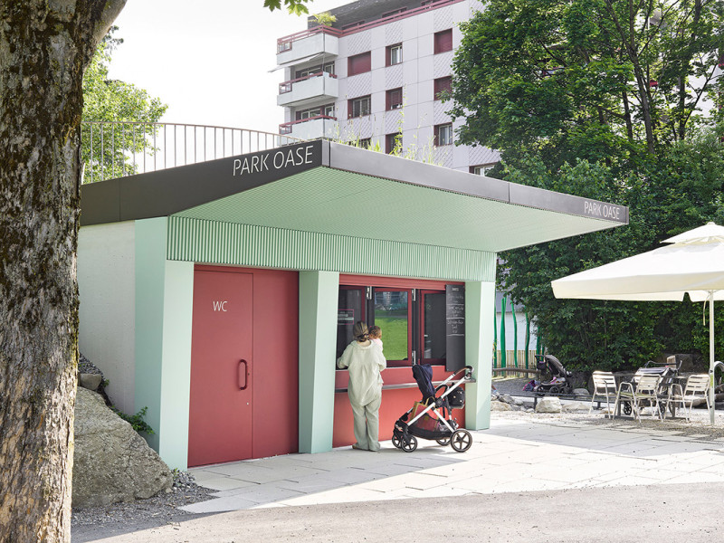 Ansicht der Buvette „Park Oase“ im Kyburgpark Sursee mit Ausschankfenster, roter Tür, grünem Vordach und angrenzendem Sitzbereich im Freien.